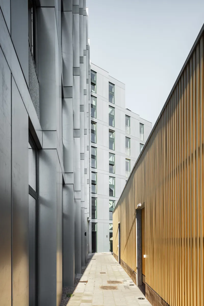 Narrow pedestrian passageway between metal panel facade and vertical timber slat wall with a figure walking