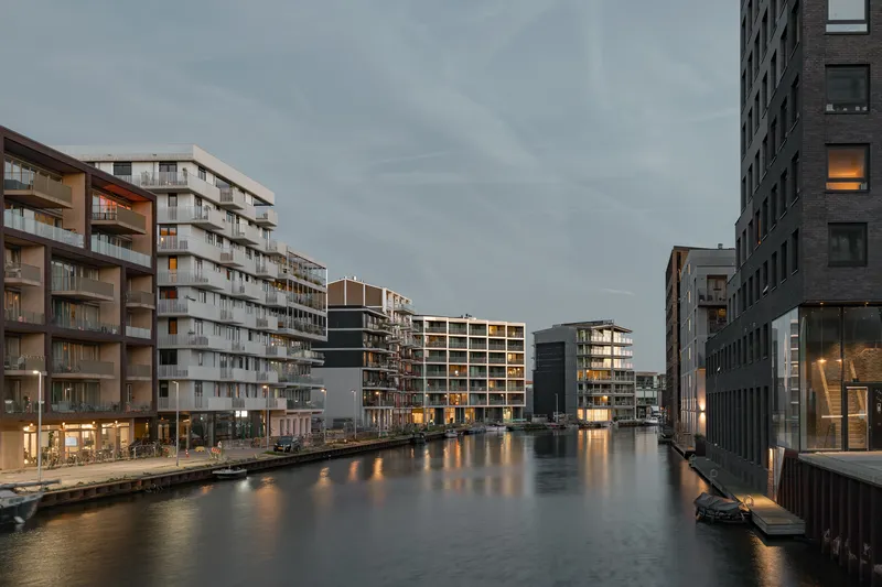 Waterside housing blocks flanking a canal at dusk with interior lights reflecting on the water