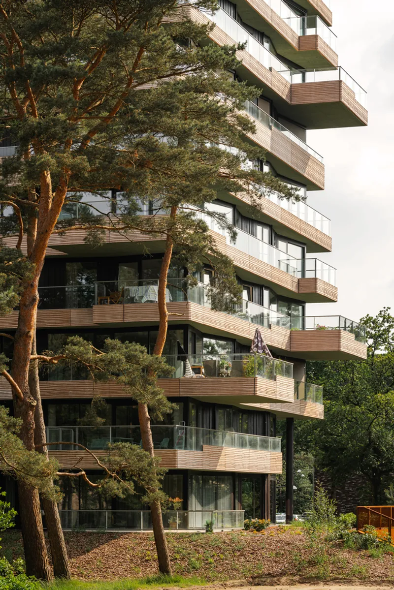 Stacked balconies with horizontal timber cladding and glass railings framed by mature pine trees