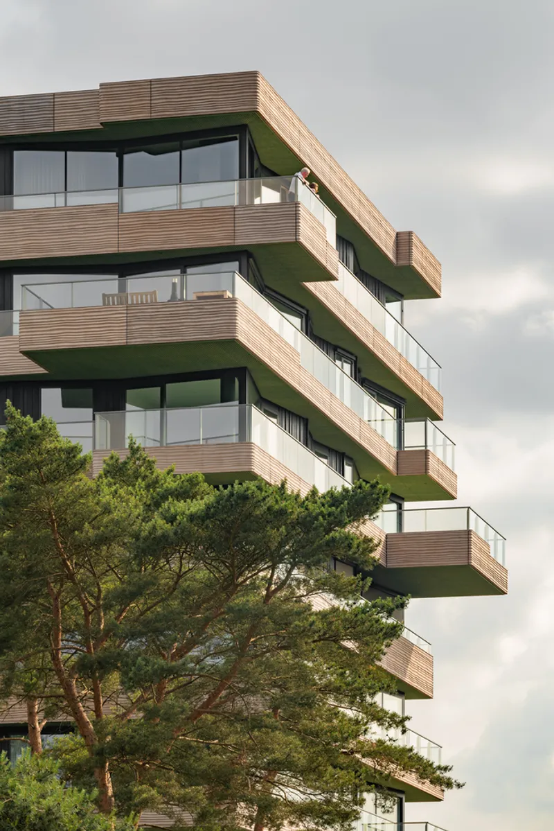 Stacked balconies with alternating timber slat facades and dark green soffits framed by a mature pine tree