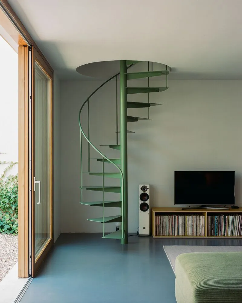 Green steel spiral staircase with open treads rising beside timber-framed glazed doors to the courtyard