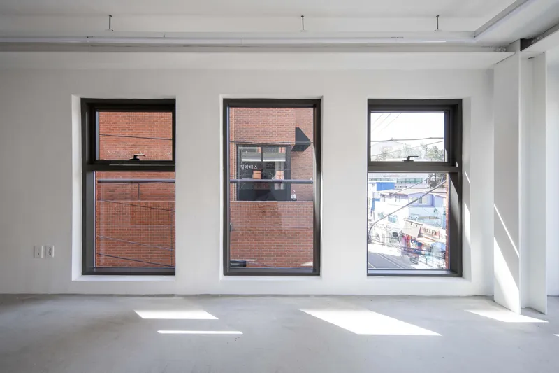 Interior view of three black-framed windows overlooking brick buildings with sunlight casting shadows on the concrete floor