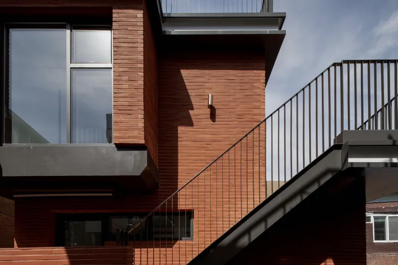 Exterior stair with metal railing ascending between red brick clad volumes under afternoon sun