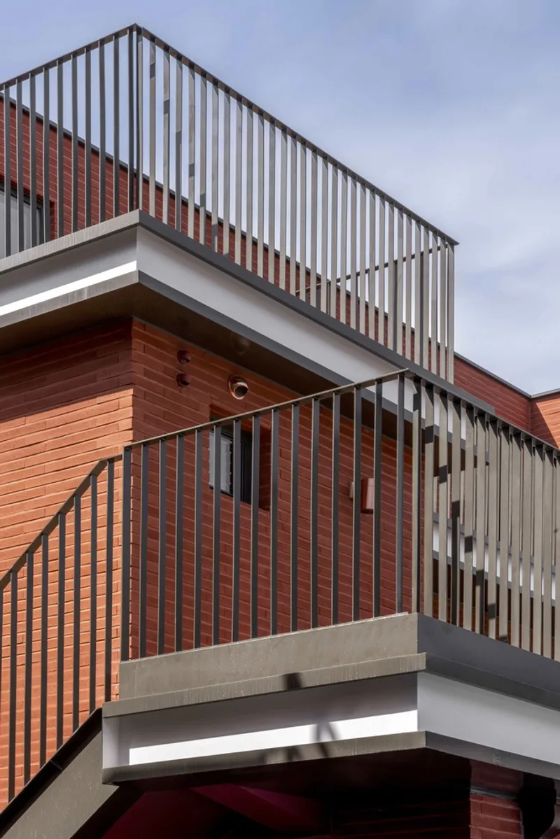Stacked balconies with vertical metal railings and red brick cladding under a cloudy sky
