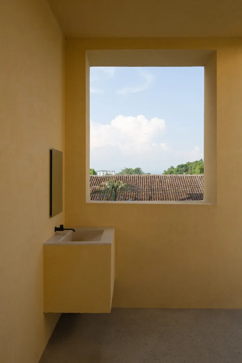 Yellow plastered bathroom with square window framing clouds and tiled rooftops beyond a floating sink