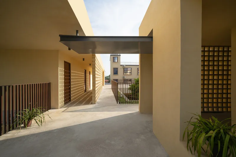 Covered walkway with perforated screen panels and overhead bridge connecting ochre plastered volumes