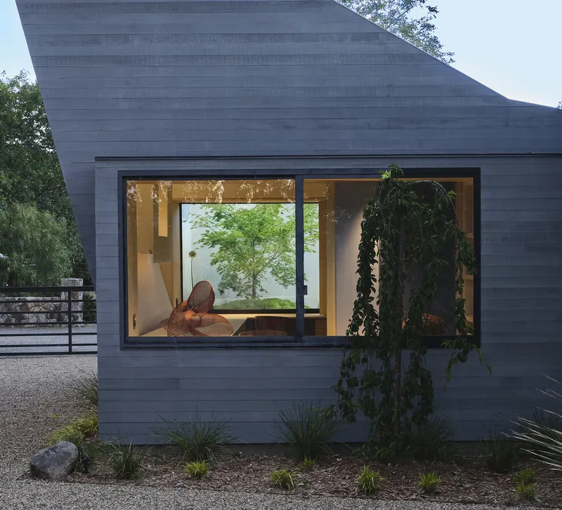 Grey horizontal timber facade with corner window revealing interior seating and courtyard maple tree at dusk