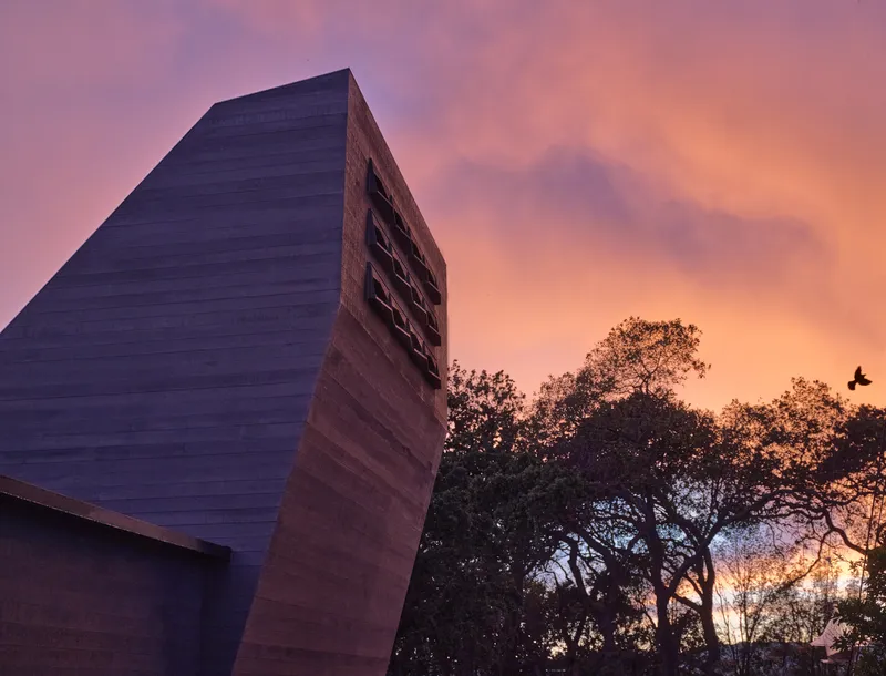 Angled tower clad in horizontal timber siding with gridded openings against a pink and orange sunset