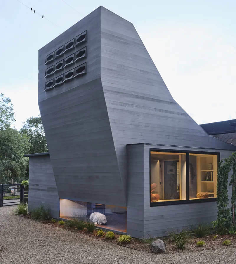 Angled concrete tower with hexagonal window openings rising above low-slung glazed volume at dusk