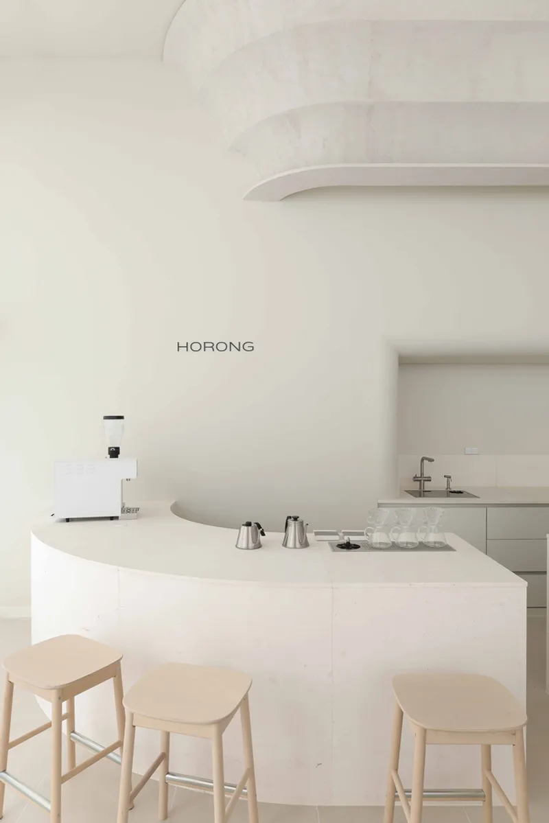 Curved white counter with coffee brewing equipment and timber stools beneath a tiered column capital