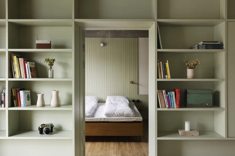 View through pale green built-in shelving toward a bedroom doorway with vertical-paneled walls