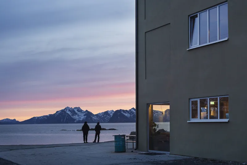Dark grey facade with glazed door and windows overlooking two figures at dusk by the sea