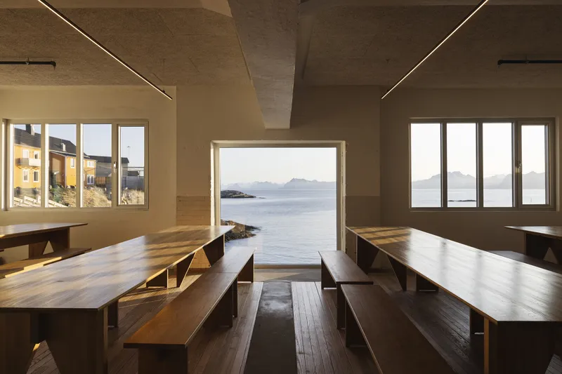 Interior hall with timber benches and tables framing a central doorway opening to the sea
