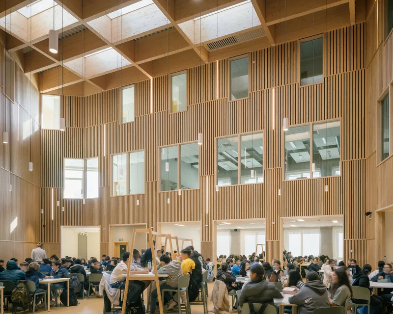 Double-height dining hall with vertical timber cladding, coffered ceiling skylights, and students at tables