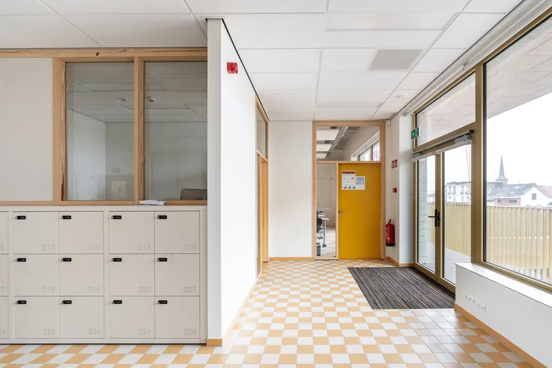 Corridor with white locker bank, checkered floor tiles, yellow door, and timber-framed windows on the right