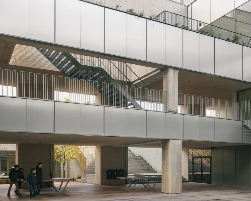 Covered entrance courtyard with concrete columns, staircases and people walking beneath a suspended terrace