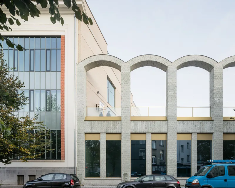 Street view of the facade with arched brick colonnade and parked cars beneath tree branches