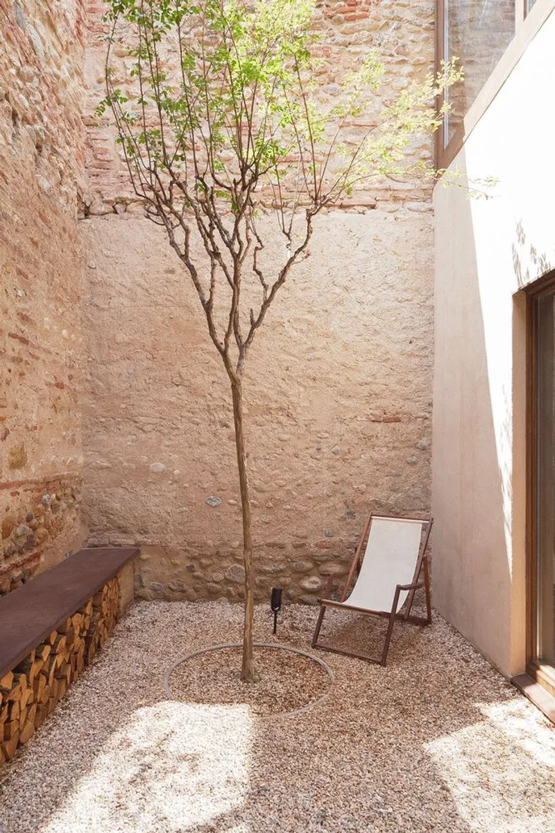 Gravel courtyard with young tree and canvas chair against a whitewashed stone wall in sunlight
