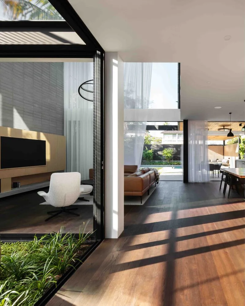 View through glass wall showing striped sunlight on timber floors and living spaces beyond the courtyard