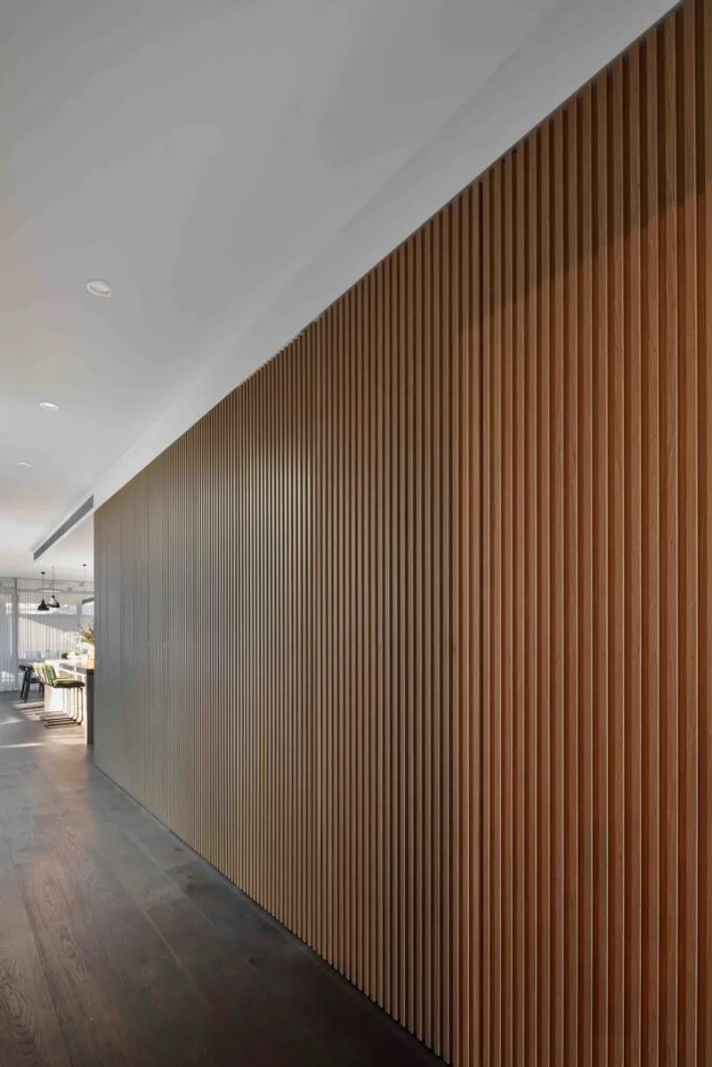 Hallway with continuous vertical timber slat partition wall under a sloped white ceiling and recessed lighting