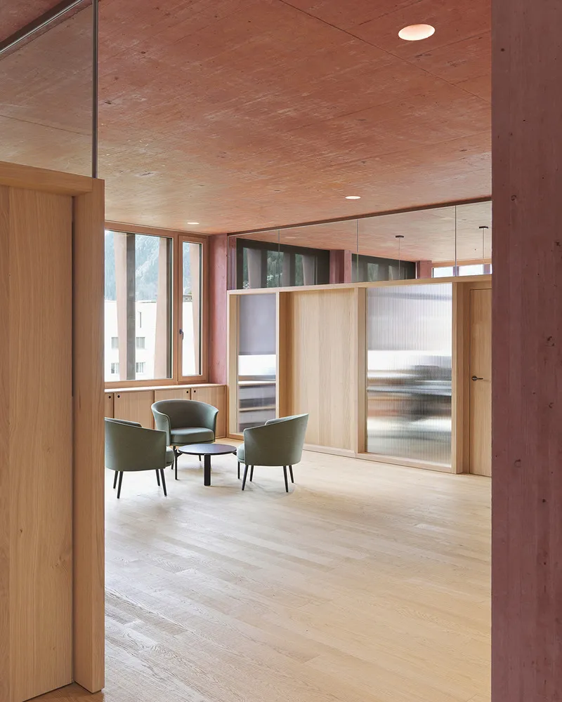 Interior lounge nook with light wood storage units, green upholstered chairs, and courtyard windows