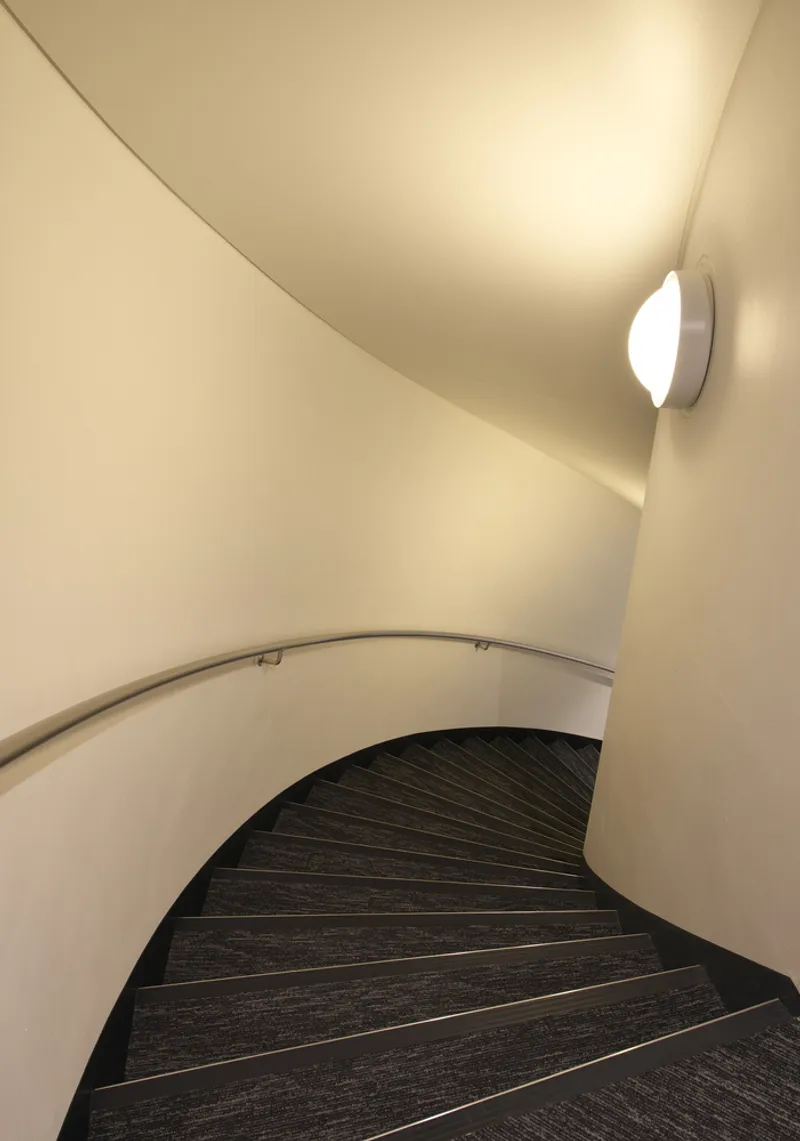 Curved stairwell with white walls, circular handrail, and wall-mounted sconce casting shadow