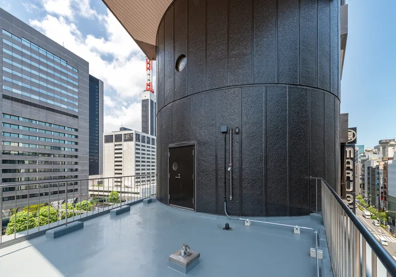 Rooftop terrace with cylindrical charcoal-paneled volume, metal door, and surrounding high-rise buildings under cloudy sky