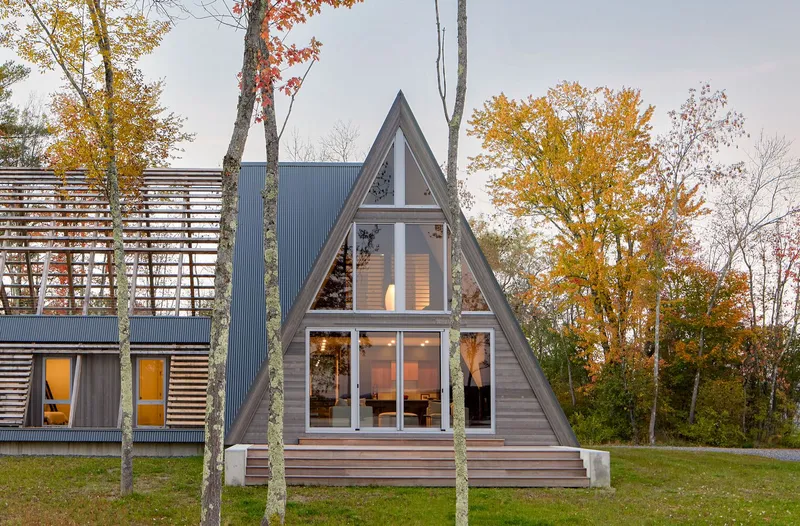 Triangular gabled volume with horizontal wood siding and full-height glazing among autumn trees at dusk