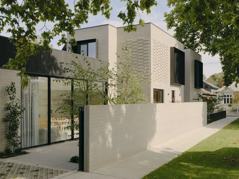 Street view of the pale brick facade with textured panels and dark vertical cladding framed by overhanging trees