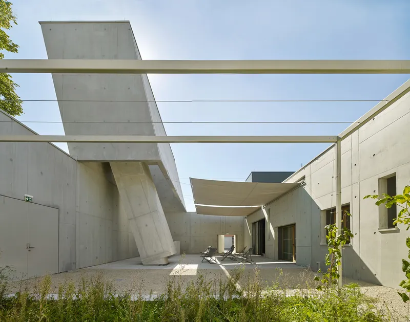 Courtyard enclosed by concrete walls with angled columns supporting horizontal canopy beams overhead in bright daylight