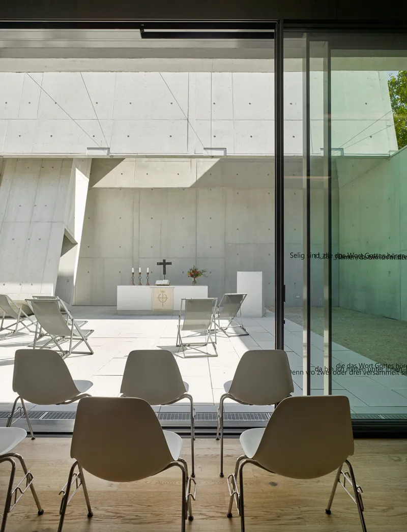 View through glass doors from interior seating toward outdoor altar framed by concrete walls and angled structure