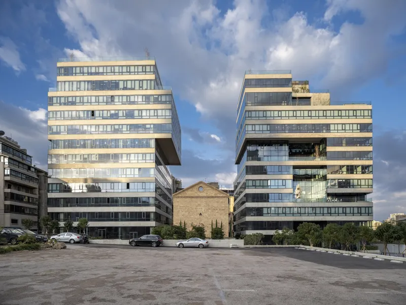 Two cantilevered glass and stone office towers flanking a small historic chapel in a paved plaza