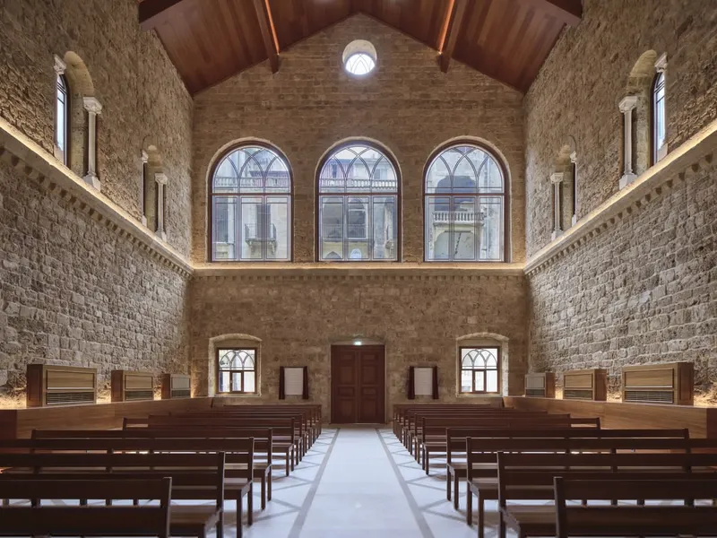 Interior nave with timber vaulted ceiling, stone walls, wooden pews and triple arched windows at the altar