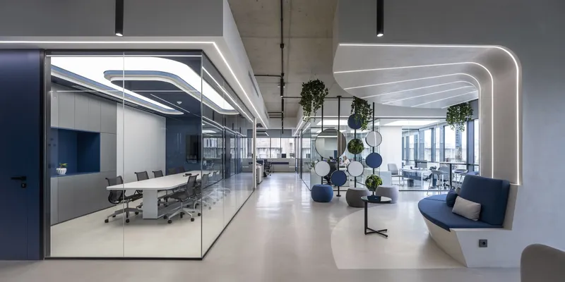 Open-plan office corridor with curved linear lighting, suspended planters, and glass-walled meeting rooms
