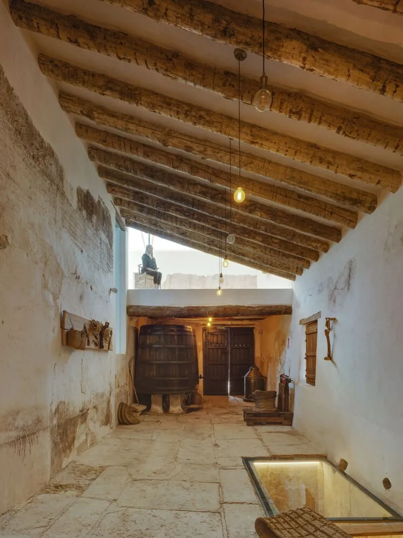 Interior passage with exposed timber ceiling beams, white plaster walls, and a wooden barrel beside historic tools