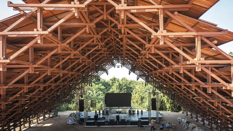Interior view through exposed timber truss framework toward a stage with trees beyond