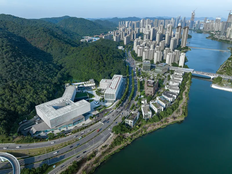 Drone view showing the complex positioned between forested hills and a curving river with highway infrastructure