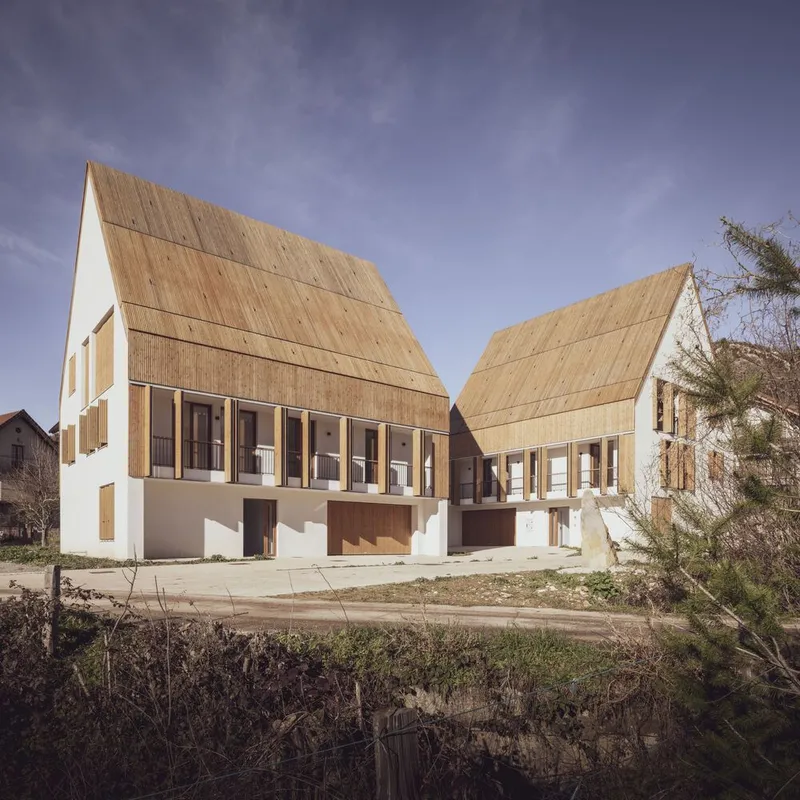 Pair of gabled timber-roofed volumes with white plaster bases and rows of vertical shutters at dusk