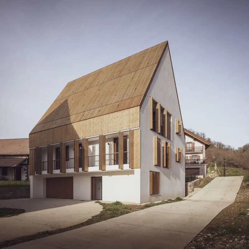 Street view of the gabled timber-clad facade with white plaster base and vertical timber shutters