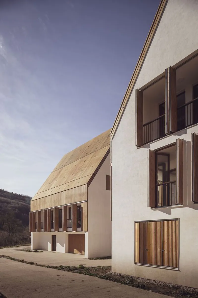 Two gabled volumes with white plaster walls and vertical timber shutters on a sloped paved street