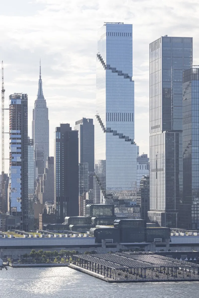 Glass tower with diagonal stepping pattern rising among neighboring highrises under overcast sky