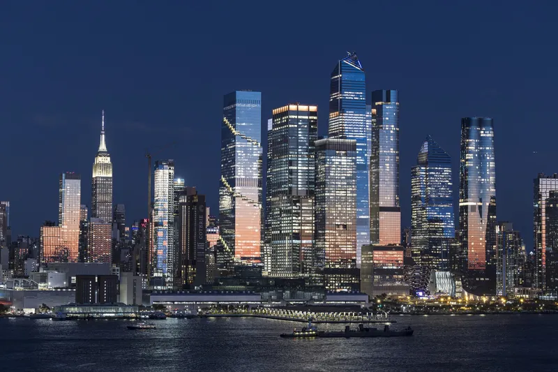 Cluster of illuminated glass towers rising above the waterfront at night with boats passing below