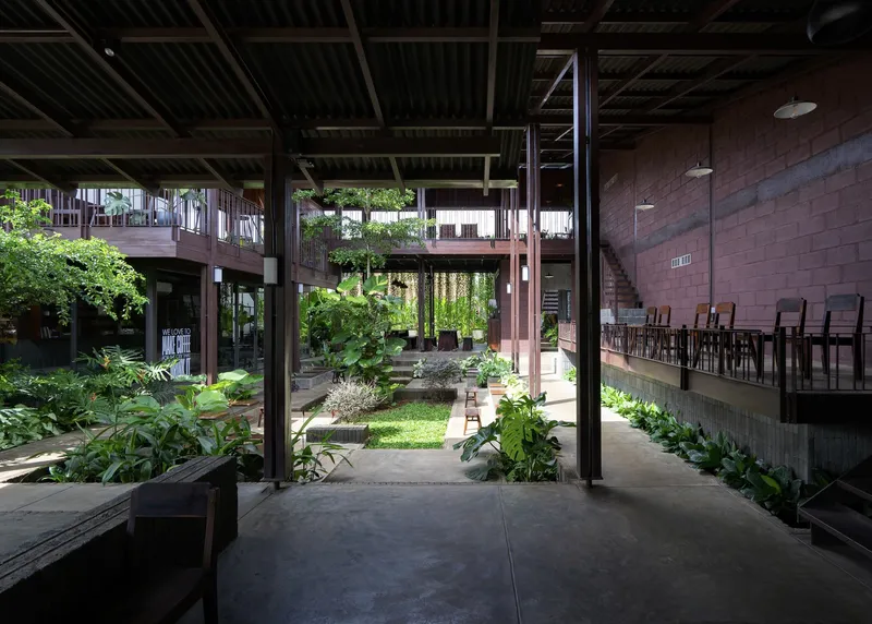 View through the covered walkway toward a sunlit courtyard with lawn, planted beds, and steel columns