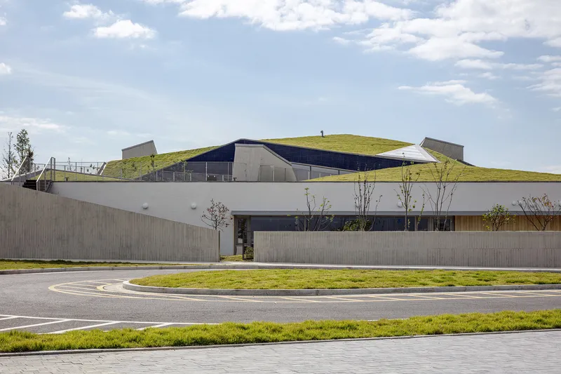 Concrete perimeter wall and grass-covered roof rising above the curved roadway under scattered clouds