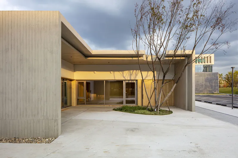 Covered entry courtyard with concrete paving and young trees surrounded by ribbed concrete walls at dusk