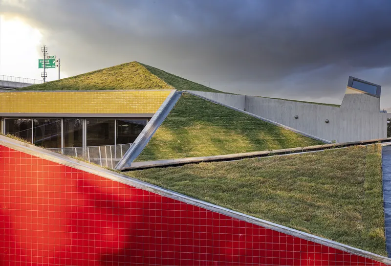 Red tile facade meeting the grass-covered roof planes under an overcast sky with storm clouds