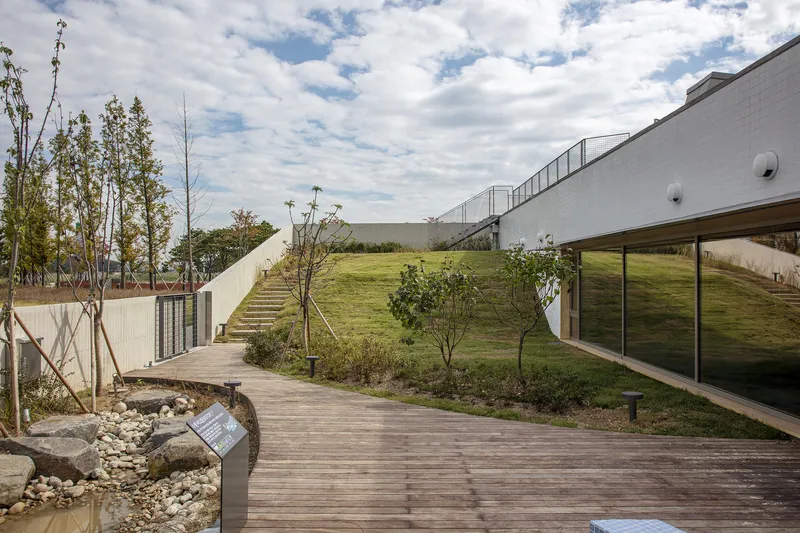 Timber boardwalk curving past a planted hillside with stairs ascending to a building above
