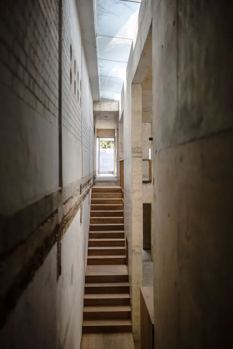 Timber staircase ascending through a narrow concrete passage illuminated by an overhead skylight