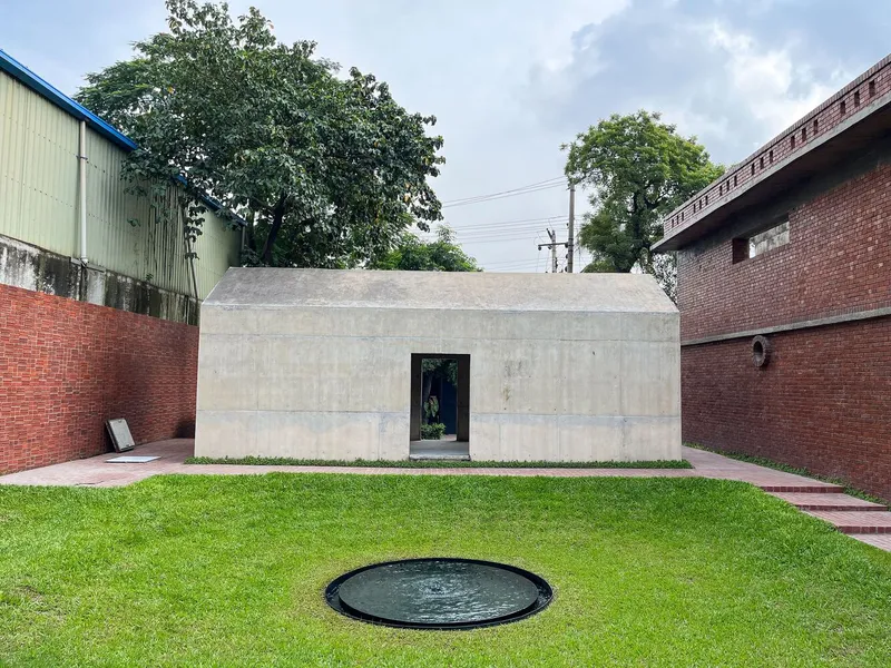 Concrete pavilion with single doorway in a courtyard bordered by brick walls and mature trees