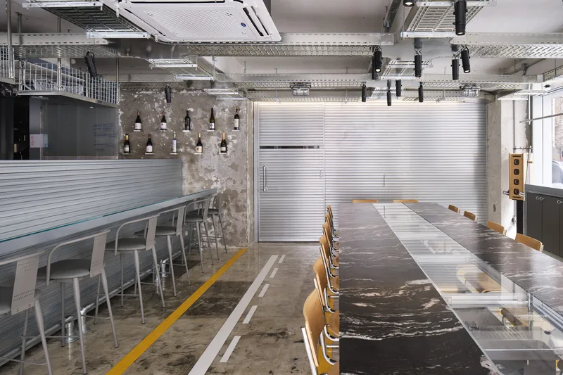 Interior dining space with exposed concrete columns, corrugated metal partitions, and a long communal table with wooden chairs